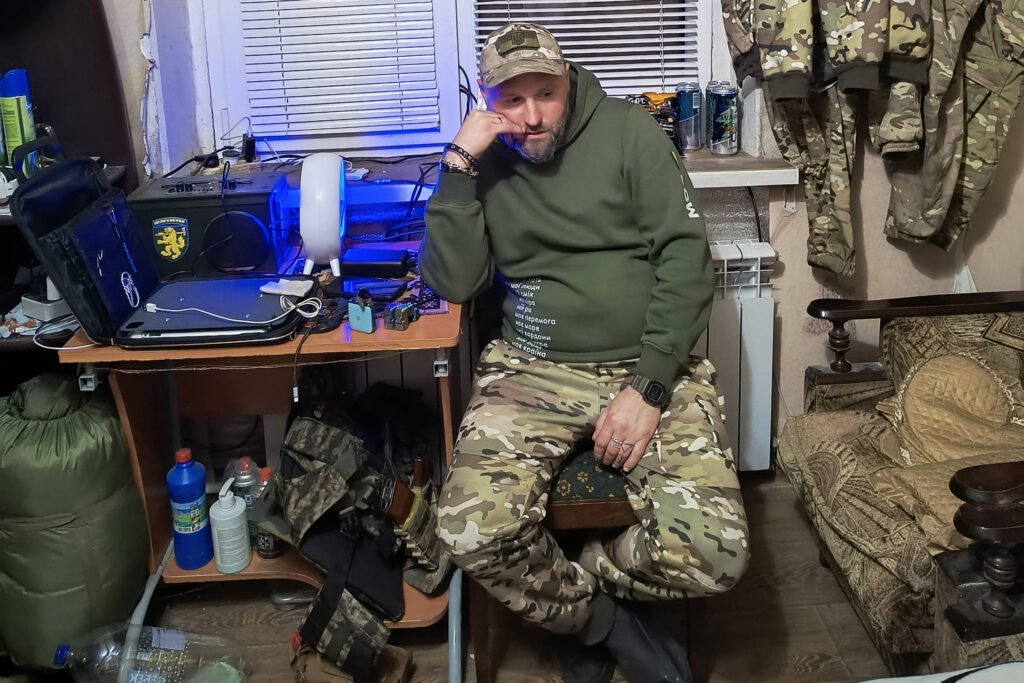Um homem sentado em uma cadeira em um ambiente com decoração militar. Ele usa um moletom verde e calças camufladas. Atrás dele, há uma bandeira da Ucrânia com um desenho de um esqueleto e um soldado. O ambiente contém uma mesa com equipamentos eletrônicos, como um computador, e várias peças de roupa militar penduradas nas paredes.