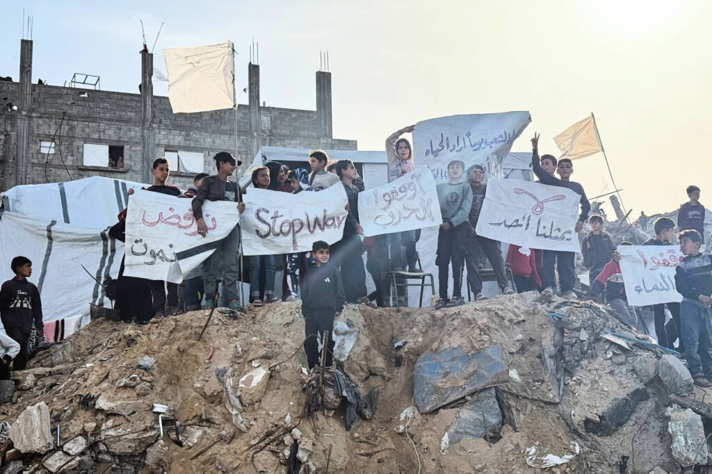 Um grupo de pessoas, incluindo crianças, está em pé sobre uma pilha de escombros, segurando cartazes. Alguns cartazes têm inscrições em árabe e um deles diz