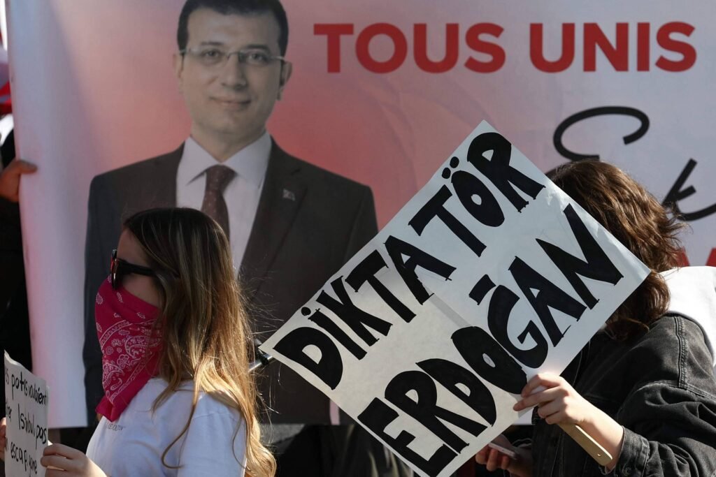 A imagem mostra um protesto com pessoas segurando cartazes. Uma mulher à esquerda, com um lenço vermelho cobrindo parte do rosto, segura um cartaz que diz