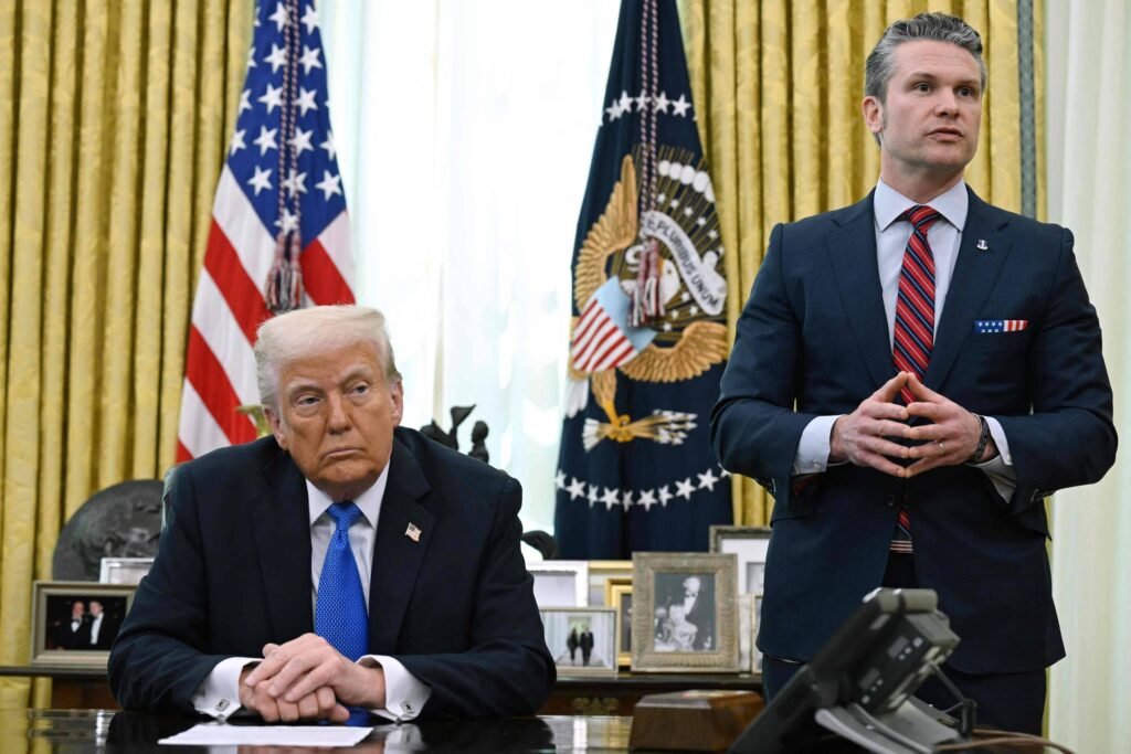 A imagem mostra Donald Trump sentado à mesa no Salão Oval, com uma expressão séria. Ele está vestido com um terno escuro e uma gravata clara. Ao fundo, há uma bandeira dos Estados Unidos e o brasão presidencial. Um segundo homem, Pete Hegseth, está em pé ao lado, com as mãos juntas, vestido com um terno escuro e uma gravata listrada. O ambiente é decorado com cortinas douradas e várias fotos emolduradas na mesa.