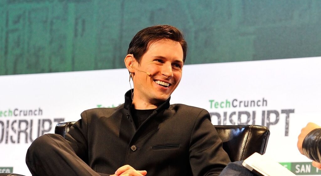 Pavel Durov com uma roupa preta, sentado no palco de um evento com um painel de fundo verde.