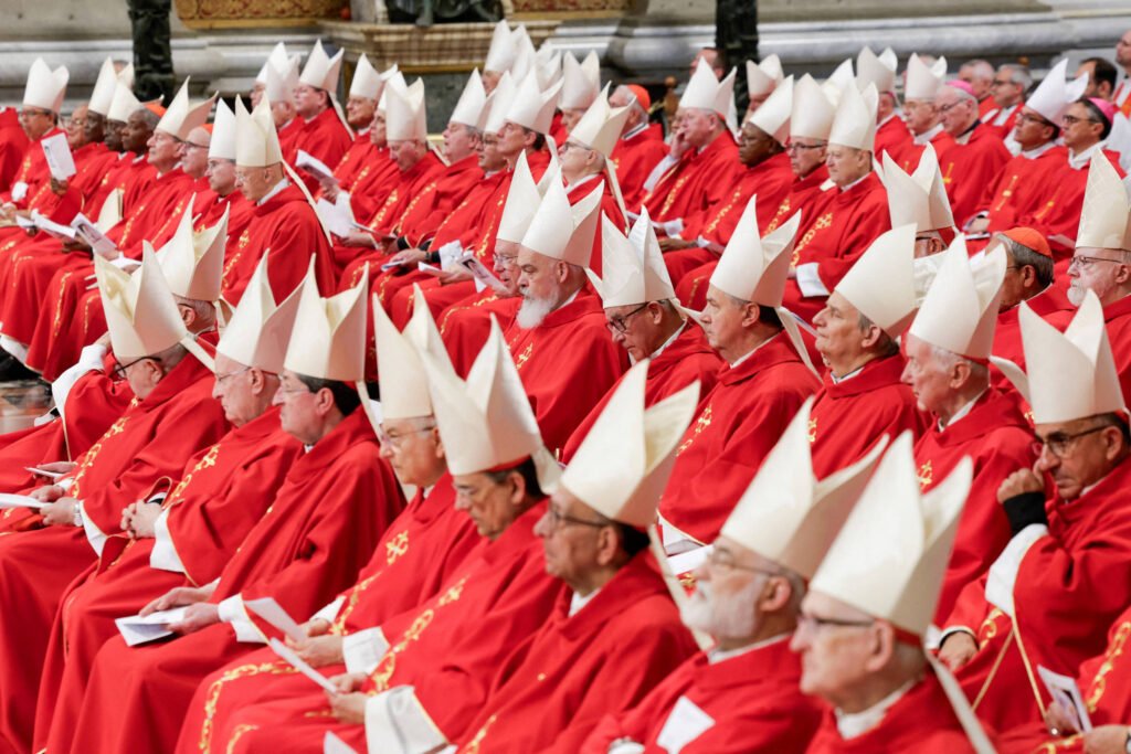 A imagem mostra um grande grupo de cardeais sentados em uma cerimônia religiosa. Eles estão vestidos com vestes vermelhas e usam mitras brancas. A maioria dos cardeais está olhando para frente, segurando papéis nas mãos. O ambiente parece ser uma catedral, com detalhes arquitetônicos visíveis ao fundo.