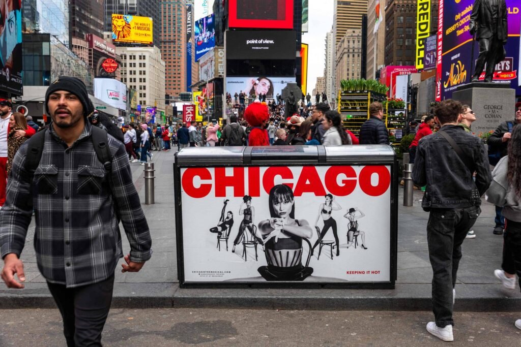 A imagem mostra uma cena movimentada em Times Square, com várias pessoas caminhando. Em primeiro plano, há um homem usando uma camisa xadrez e um boné, passando ao lado de um painel publicitário que exibe a palavra