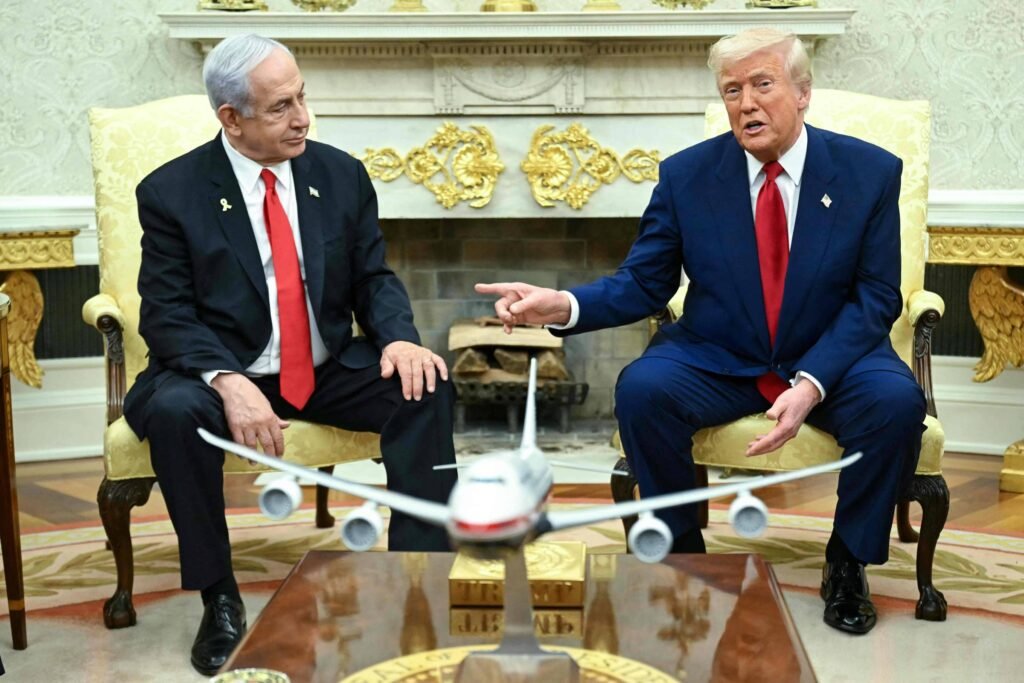 A imagem mostra duas figuras sentadas em cadeiras no Salão Oval da Casa Branca. À esquerda, um homem com cabelo grisalho e terno escuro, e à direita, um homem com cabelo loiro e terno escuro. Entre eles, há um modelo de avião em uma mesa de centro. O ambiente é decorado com um grande espelho e detalhes em dourado.