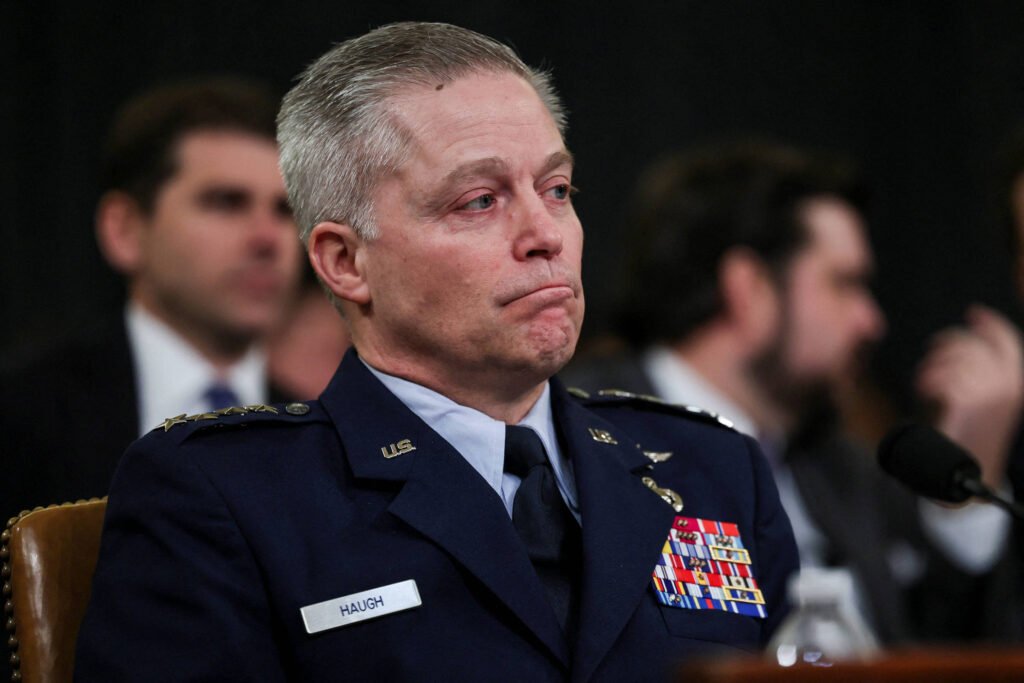 Um oficial militar, vestido com um uniforme azul escuro, está sentado em uma mesa durante uma audiência. Ele tem uma expressão séria e parece emocionado. No fundo, outras pessoas estão visíveis, algumas olhando para ele. O oficial possui várias medalhas e insígnias em seu uniforme.