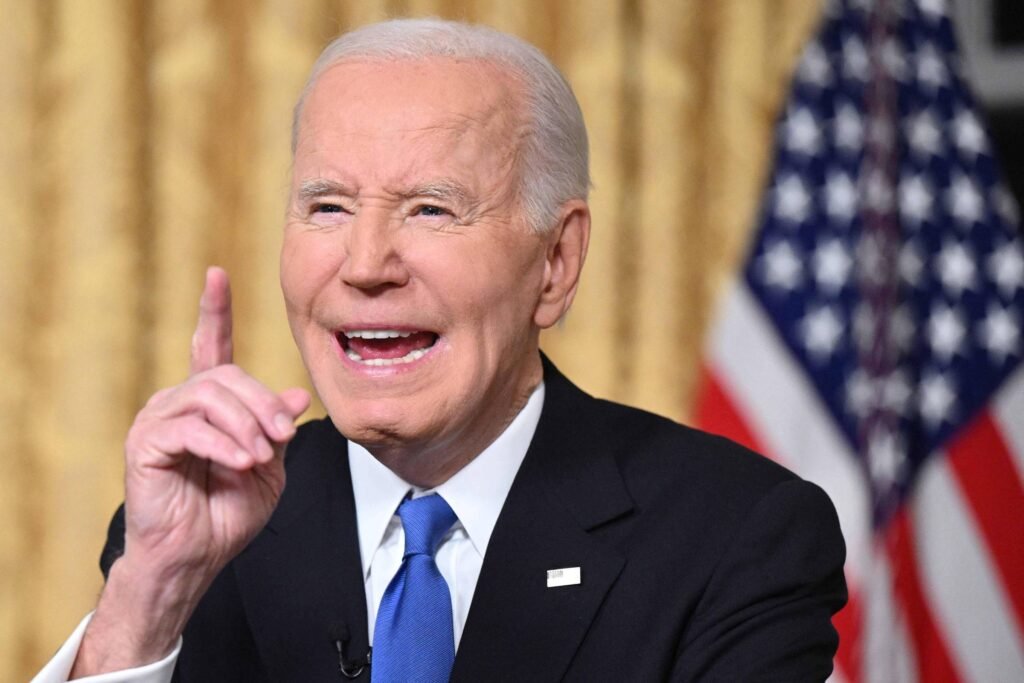 Joe Biden, um homem de cabelo grisalho, vestindo um terno escuro e uma gravata azul, está falando em um microfone. Ele tem uma expressão animada e gesticula com a mão levantada, como se estivesse enfatizando um ponto. Ao fundo, há uma cortina dourada e uma bandeira dos Estados Unidos.