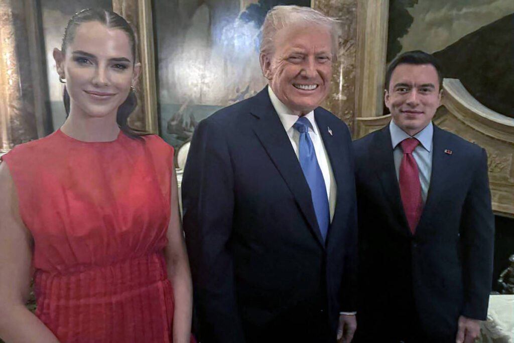 A imagem mostra três pessoas posando para uma foto em um ambiente interno elegante. À esquerda, uma mulher com cabelo preso e vestido vermelho sorri. No centro, um homem com cabelo claro e terno escuro, sorrindo amplamente. À direita, um homem com cabelo escuro, usando um terno escuro e gravata vermelha, também sorrindo. Ao fundo, há paredes decoradas com pinturas.