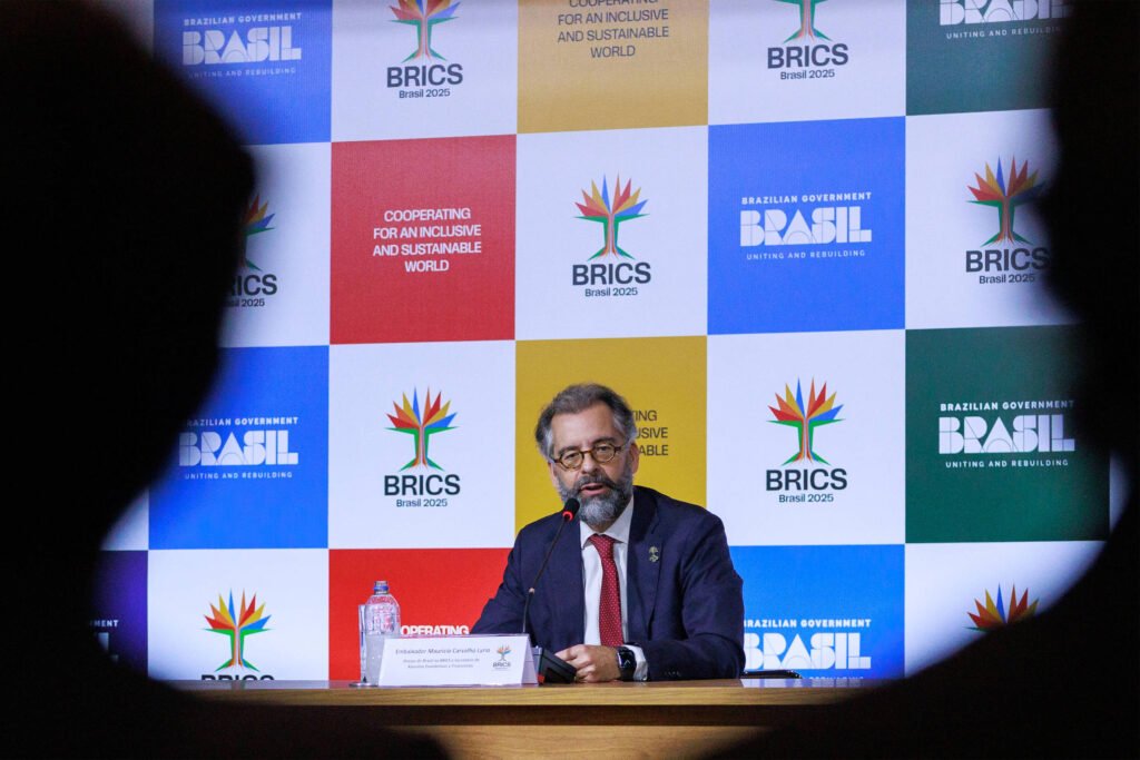 Um homem com barba e óculos está sentado em uma mesa, falando em um microfone. Ele usa um paletó escuro e uma camisa clara. Ao fundo, há um painel colorido com logotipos do BRICS e a frase