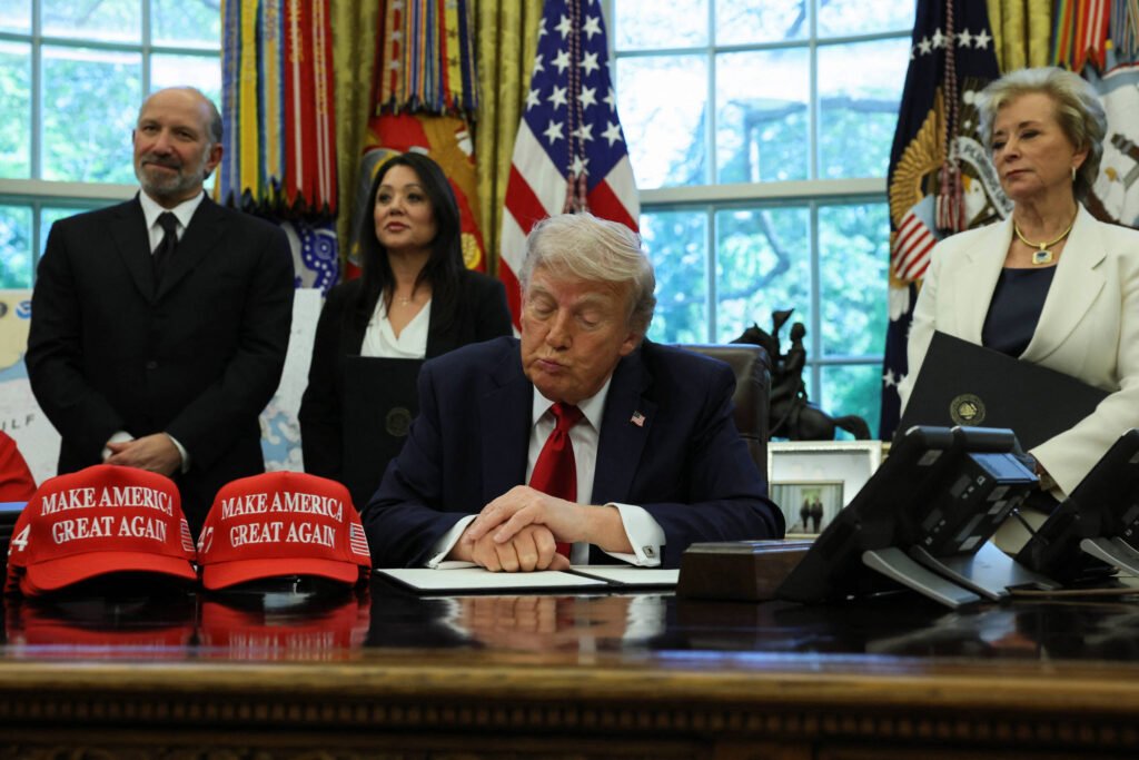 A imagem mostra um grupo de pessoas no Salão Oval da Casa Branca. Trump, que está sentado à mesa, está assinando um documento. Ele está usando um terno escuro e uma gravata vermelha. À sua frente, há três bonés vermelhos com a frase