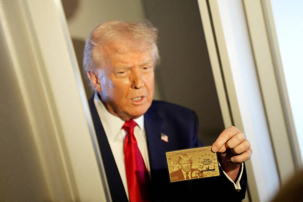 Trump, um homem com cabelo loiro e terno escuro, está segurando um cartão dourado. Ele parece estar em uma porta, com uma expressão de seriedade. O fundo é desfocado, mas parece ser um ambiente interno.