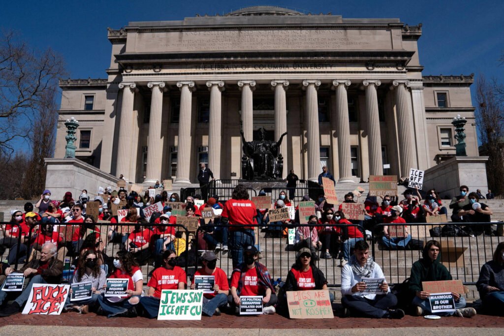 Um grupo de pessoas sentadas em frente a um grande edifício com colunas, participando de um protesto. A maioria das pessoas está vestindo camisetas vermelhas e segurando cartazes com mensagens de protesto. O ambiente é ensolarado e há uma escadaria que leva à entrada do edifício. Algumas pessoas estão usando máscaras.