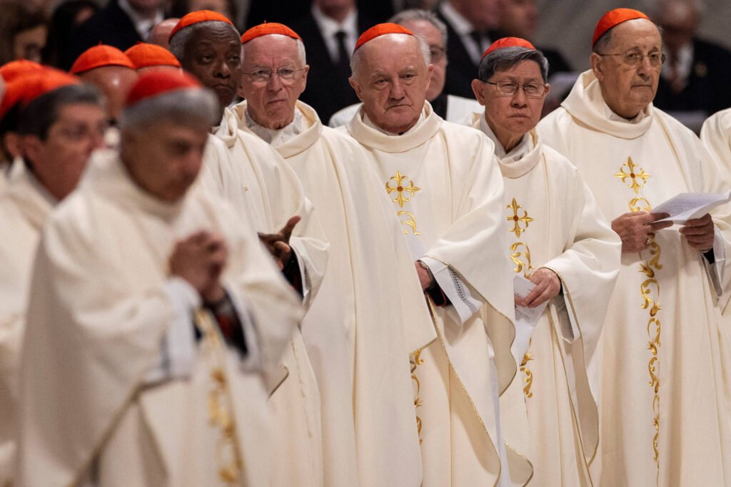 Um grupo de cardeais está reunido em uma cerimônia religiosa. Eles estão vestidos com vestes brancas e capas, e muitos usam chapéus vermelhos. O ambiente parece ser uma igreja ou catedral, com um fundo desfocado que sugere um evento solene. Os cardeais estão em diferentes posturas, alguns com as mãos unidas em oração, enquanto outros seguram papéis.