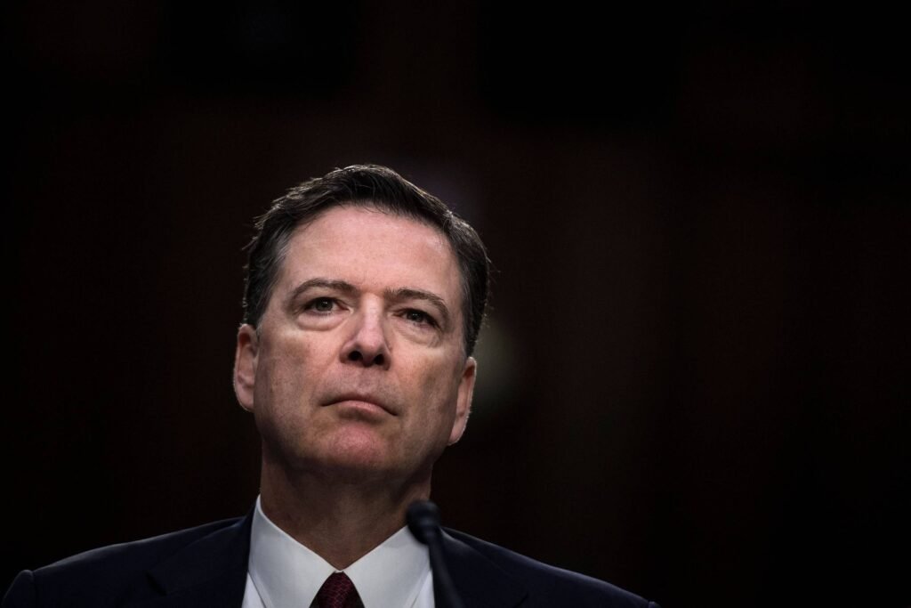 James Comey, um homem com cabelo escuro e liso, vestindo um terno escuro e uma gravata, está em um ambiente com pouca iluminação, olhando para frente com uma expressão séria. O fundo é desfocado, sugerindo um cenário formal, possivelmente uma audiência ou uma sessão de perguntas.