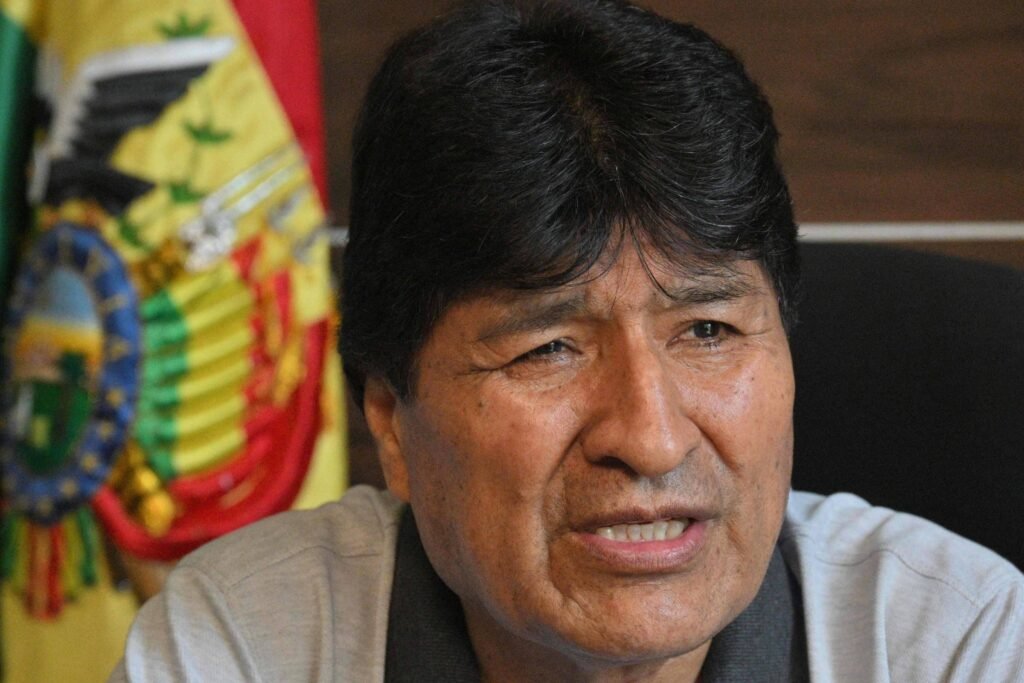 Um homem com cabelo escuro e liso, usando uma camisa cinza, está falando em um ambiente interno. Ao fundo, há uma bandeira da Bolívia, que apresenta as cores verde, amarelo e vermelho, além de um brasão. O homem parece estar expressando uma opinião ou fazendo uma declaração.
