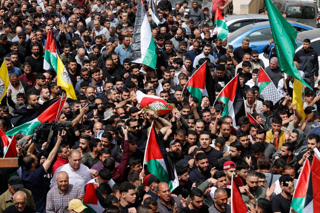 Uma grande multidão de pessoas se reúne em uma área urbana, segurando bandeiras palestinas e outras bandeiras. No centro da imagem, um grupo de pessoas carrega um caixão coberto com uma bandeira, enquanto outras pessoas ao redor demonstram apoio. A cena é marcada por uma atmosfera de luto e protesto, com muitos rostos visíveis entre a multidão.