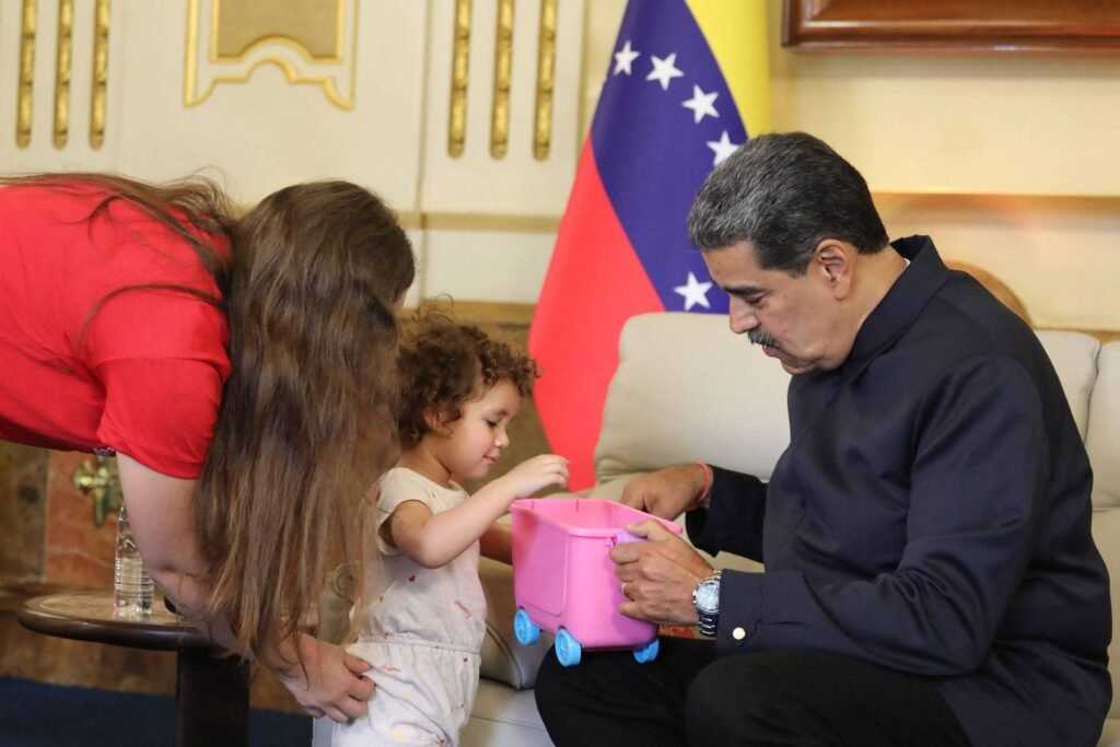 A imagem mostra um homem sentado em um sofá, olhando para uma criança que está em pé ao seu lado. A criança, com cabelo cacheado, está segurando um balde rosa. Uma mulher com cabelo longo e solto está se inclinando em direção à criança. Ao fundo, há uma bandeira da Venezuela visível. O ambiente parece ser um escritório ou sala de estar decorada.