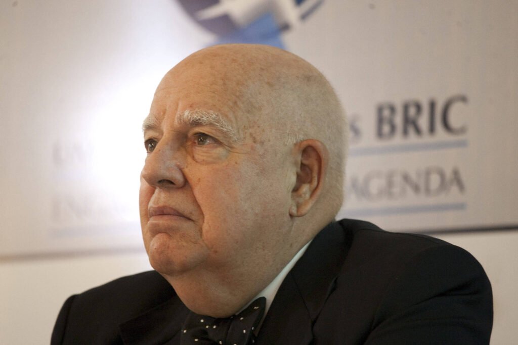 Um homem idoso com cabelo calvo e pele clara, usando um terno escuro e uma gravata borboleta. Ele está olhando para o lado, com uma expressão séria. Ao fundo, há um painel com texto que menciona