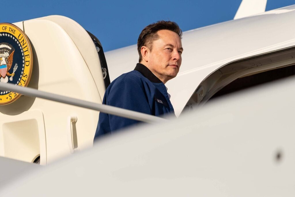 Elon Musk está em uma aeronave, com um emblema do governo dos Estados Unidos visível ao fundo. Ele usa uma jaqueta azul e parece estar olhando para fora. O céu está claro e azul, e a parte da aeronave é visível na imagem.