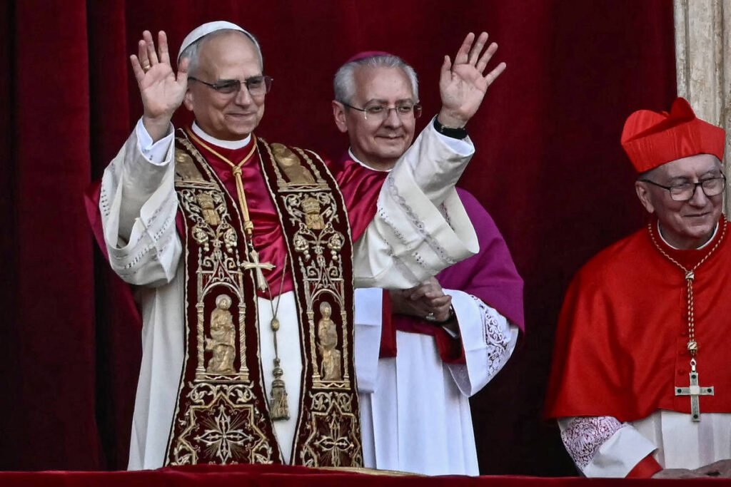 A imagem mostra um homem vestido com vestes papais, levantando as mãos em saudação. Ele está acompanhado por dois outros homens, um deles vestido com uma túnica roxa e o outro com vestes vermelhas e um chapéu cardinalício. O fundo é de um tom vermelho, sugerindo um ambiente formal, possivelmente uma cerimônia religiosa.