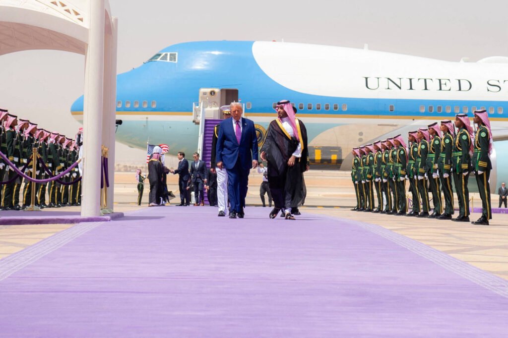 A imagem mostra uma cerimônia de recepção oficial em um aeroporto. Um homem de terno azul caminha ao lado de um homem vestido com traje tradicional, ambos se dirigindo para frente em um tapete roxo. Ao fundo, um grande avião com a inscrição