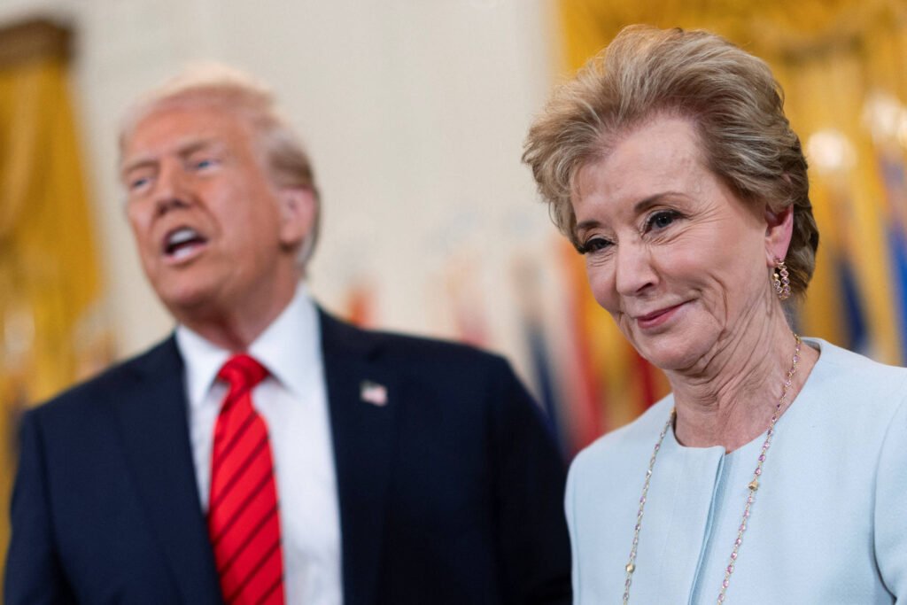 A imagem mostra um evento no Salão Oval da Casa Branca, com Donald Trump, um homem de terno escuro e gravata vermelha à esquerda, falando. À direita, Linda McMahon, uma mulher com cabelo claro e vestido azul claro sorri enquanto observa. O fundo é decorado com cortinas douradas e bandeiras.