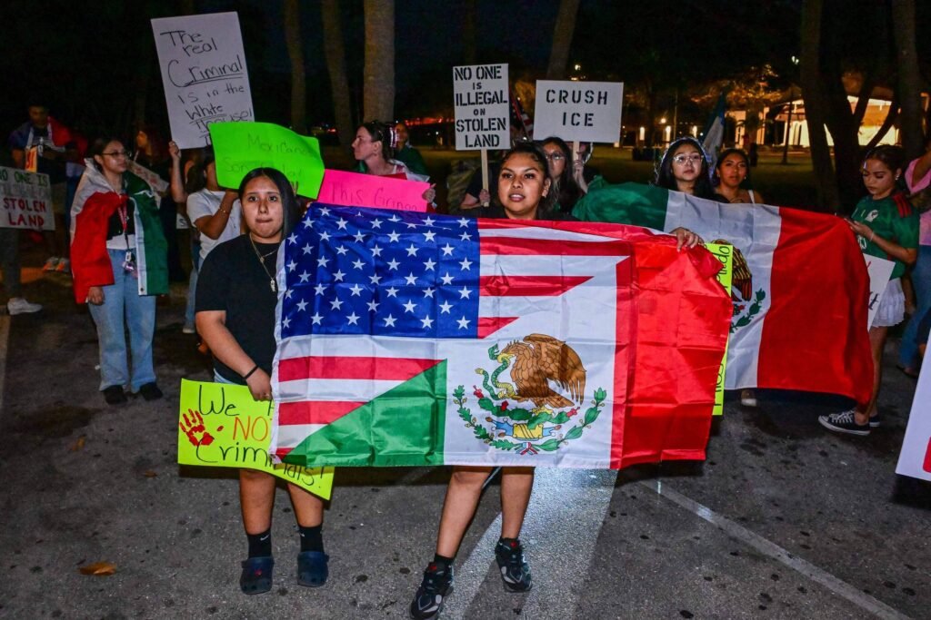 Um grupo de pessoas participa de um protesto à noite, segurando cartazes e bandeiras. No centro, duas jovens seguram uma bandeira que combina elementos dos Estados Unidos e do México. Os cartazes têm mensagens como