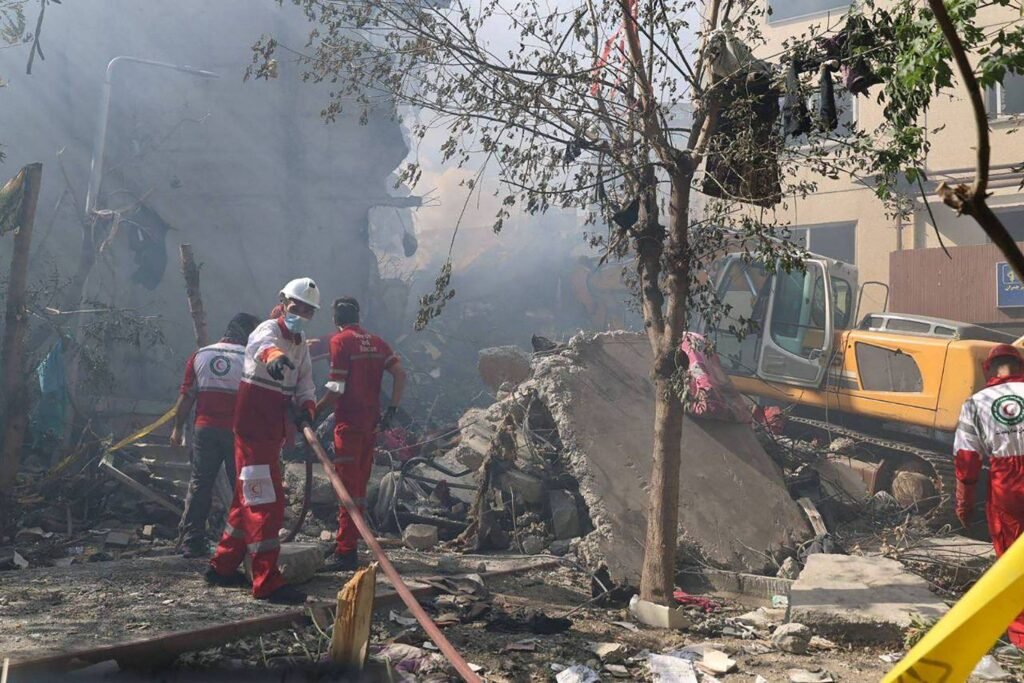 A imagem mostra um cenário de desastre, possivelmente após uma explosão ou colapso de edifício. Há fumaça no ar e escombros visíveis no chão. Vários socorristas, vestidos com uniformes vermelhos e coletes, estão trabalhando na área, alguns deles usando capacetes. Um veículo de resgate amarelo está presente ao fundo, e há uma árvore próxima aos escombros.
