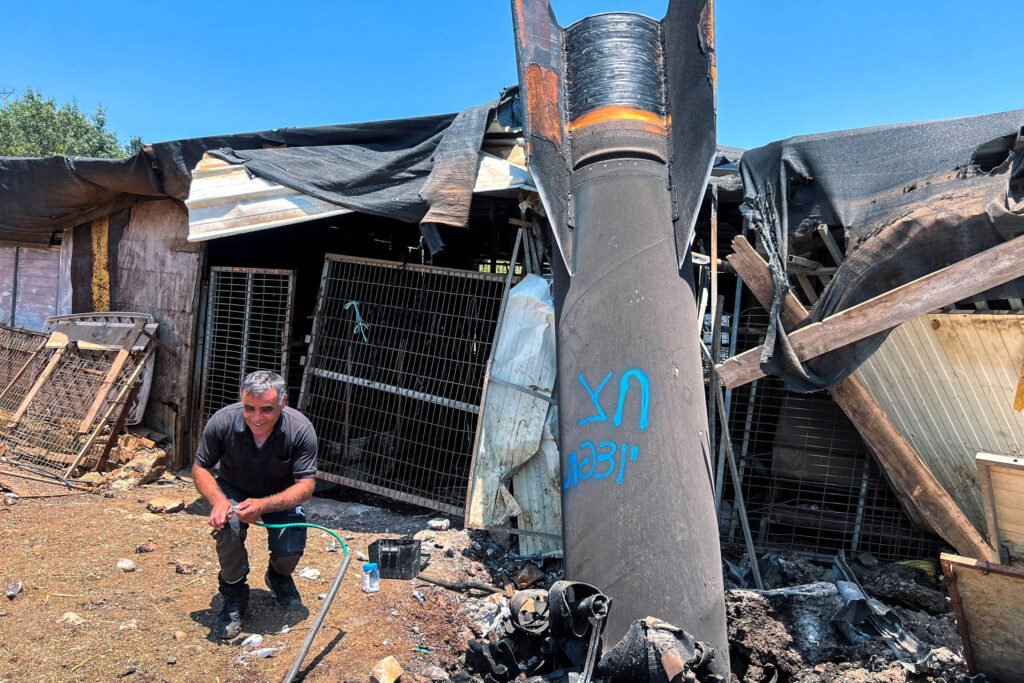 A imagem mostra um homem agachado em frente a uma estrutura danificada, que parece ser uma casa ou abrigo. Ao lado dele, há um foguete grande e danificado, com inscrições azuis visíveis em sua superfície. O fundo da imagem revela uma construção em ruínas, com telhados rasgados e materiais espalhados pelo chão, sugerindo um cenário de destruição. O céu está claro e ensolarado, contrastando com a gravidade da cena.