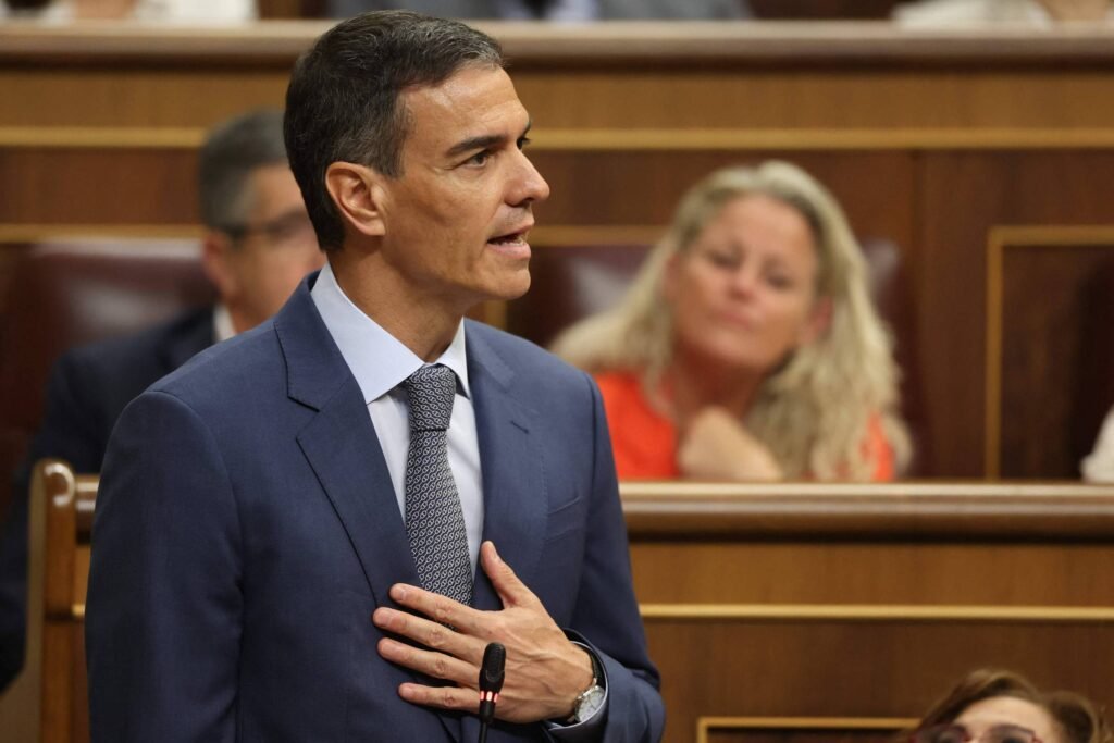 Um homem em um terno azul está falando em um microfone em um ambiente de parlamento. Ele tem cabelo curto e está gesticulando com a mão sobre o coração. Ao fundo, há várias pessoas, incluindo uma mulher com cabelo longo e loiro, que está olhando para ele. O ambiente é decorado com painéis de madeira e há cadeiras em estilo formal.