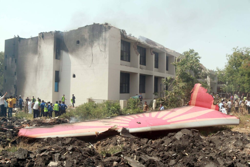 A imagem mostra os destroços de um avião que colidiu com um edifício. O avião está parcialmente destruído, com uma das asas visível no chão. O edifício ao fundo apresenta danos significativos, com janelas quebradas e fumaça saindo do telhado. Há várias pessoas ao redor, algumas observando e outras possivelmente ajudando. A cena é de caos e destruição, com vegetação ao redor.