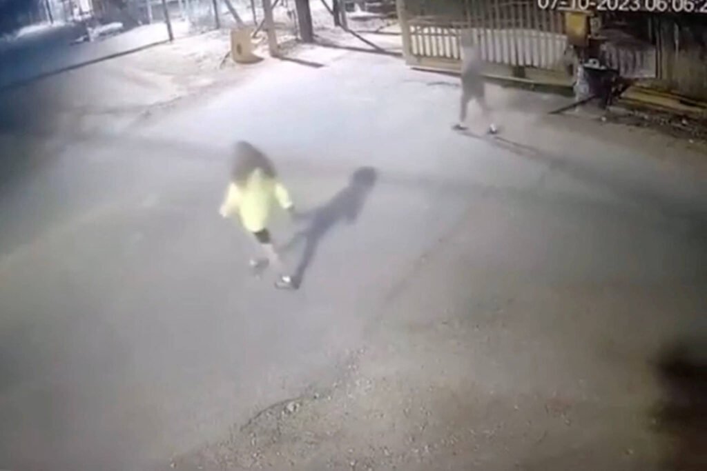 A imagem mostra uma cena noturna capturada por uma câmera de segurança. No centro, uma pessoa com um casaco amarelo caminha em direção à câmera, enquanto outra figura, vestindo roupas escuras, se move ao fundo, perto de um portão. O ambiente parece ser uma rua ou área externa, com iluminação fraca e algumas sombras visíveis.