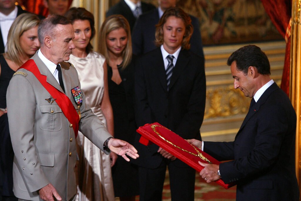 A imagem mostra uma cerimônia de condecoração, onde um homem em uniforme militar, com uma faixa vermelha, está recebendo uma medalha de um homem em terno. Ao fundo, há várias pessoas observando, incluindo mulheres em vestidos e um homem com gravata. O ambiente parece ser um salão decorado, possivelmente em um palácio.