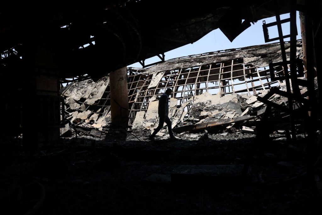 A imagem mostra uma pessoa caminhando entre os escombros de um edifício destruído por um incêndio. O ambiente é escuro e carbonizado, com estruturas metálicas expostas e janelas quebradas ao fundo. A luz do sol entra pela abertura, iluminando parcialmente a cena.