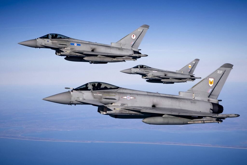 A imagem mostra três caças Eurofighter Typhoon voando em formação no céu. Os aviões são de cor cinza e estão em um ambiente aéreo claro, com uma vista do solo ao fundo. O caça da frente possui uma insígnia na fuselagem, enquanto os outros dois estão posicionados um acima e um ao lado do primeiro.