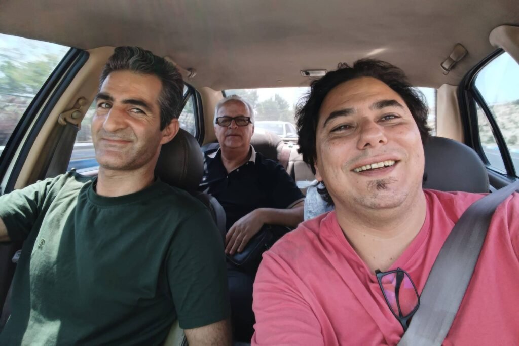 Três homens estão dentro de um carro. O homem à esquerda está dirigindo, vestindo uma camiseta verde. O homem ao centro está sentado no banco traseiro, usando óculos e uma camisa preta. O homem à direita está tirando uma selfie, vestindo uma camiseta rosa. A luz do sol entra pela janela, iluminando o interior do carro.
