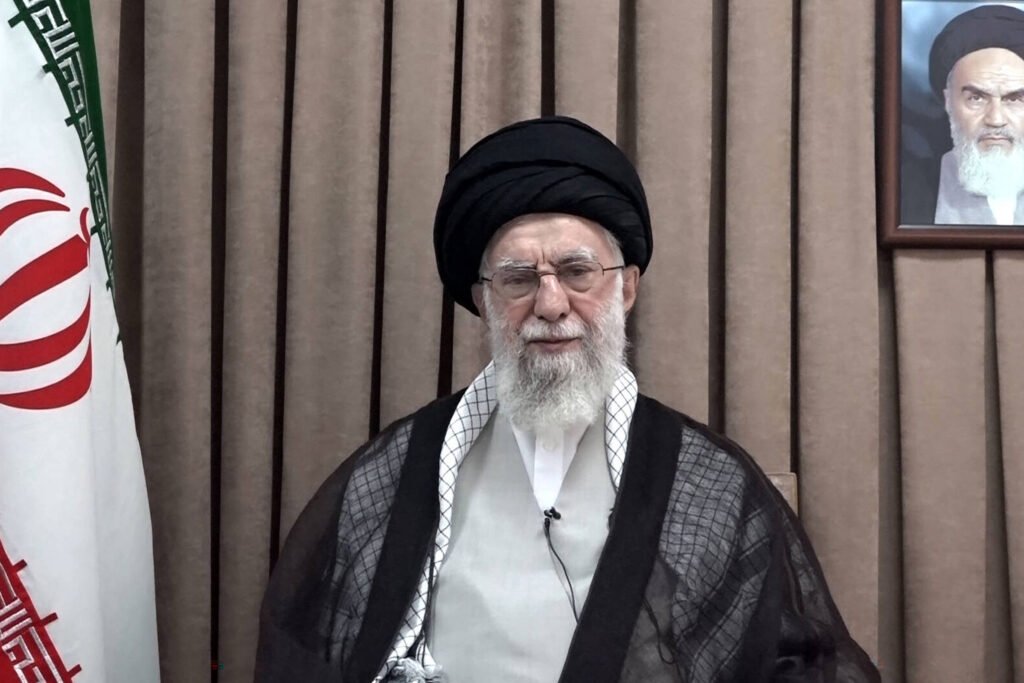 Ali Khamenei, um homem com barba e óculos, está vestido com um manto preto e uma túnica branca e sentado em frente a uma bandeira do Irã. Ao fundo, há uma parede com cortinas marrons e um retrato de outra figura religiosa na parede à direita. O homem parece estar falando ou se preparando para um discurso.