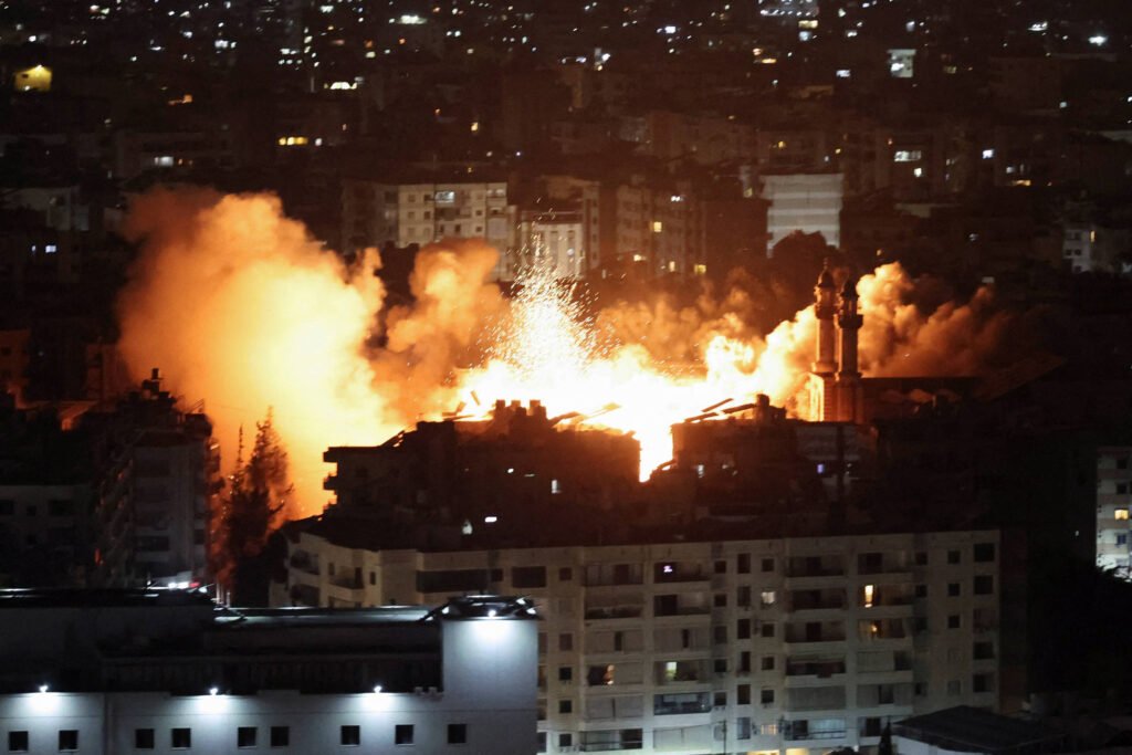 A imagem mostra uma explosão em uma área urbana durante a noite. Chamas e fumaça laranja se elevam de um edifício, iluminando a cena ao redor. Vários prédios podem ser vistos ao fundo, com luzes acesas nas janelas. O céu está escuro, e a fumaça se espalha pela atmosfera.