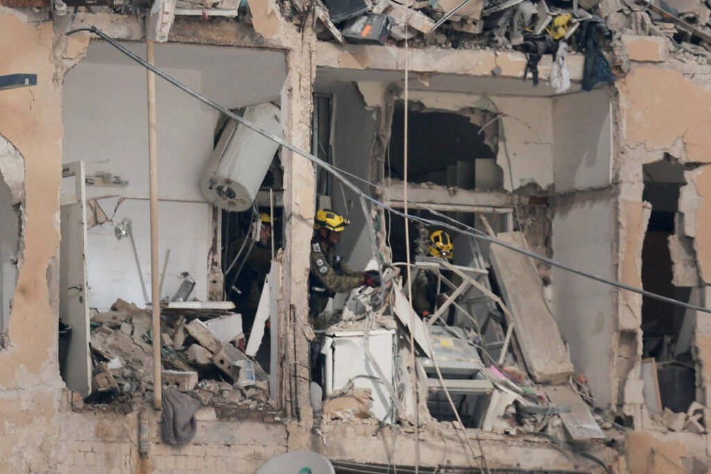 A imagem mostra um edifício severamente danificado, com várias paredes e janelas destruídas. Há escombros visíveis e partes da estrutura expostas. Dois bombeiros, usando capacetes, estão visíveis no interior do edifício, entre os destroços, aparentemente realizando operações de resgate. Fios elétricos pendem do teto e há um tanque de água visível em uma das paredes.