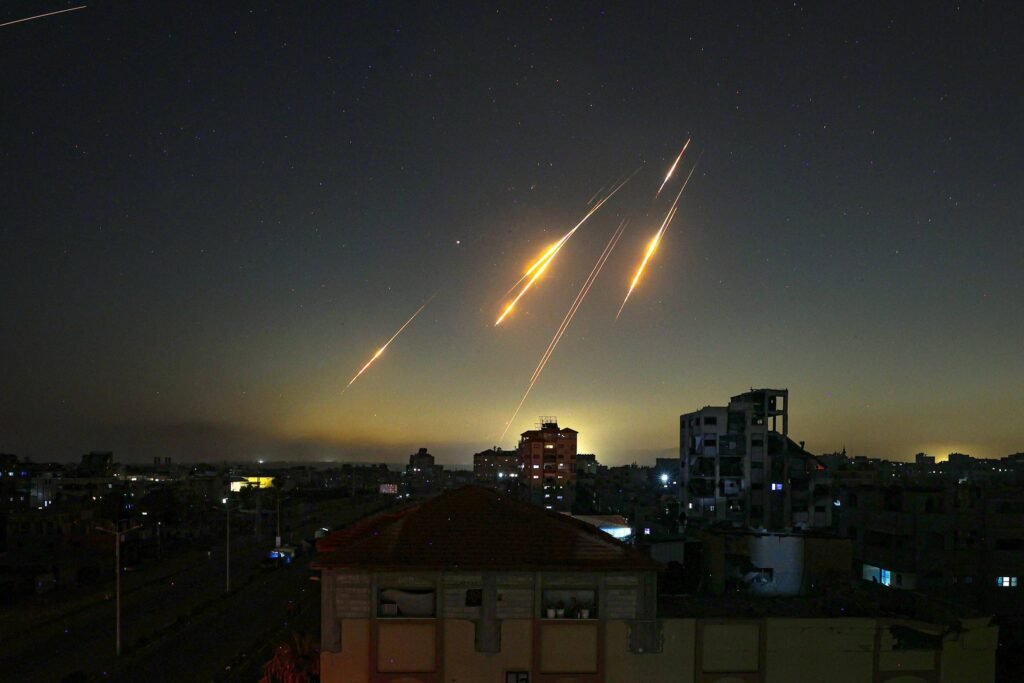 A imagem mostra um céu noturno com várias trilhas de luz, indicando o lançamento de mísseis ou foguetes. Abaixo, há uma vista de uma cidade com edifícios e ruas visíveis, enquanto o horizonte apresenta um brilho proveniente de uma fonte de luz, possivelmente uma explosão ou incêndio. O céu é escuro, com algumas estrelas visíveis.