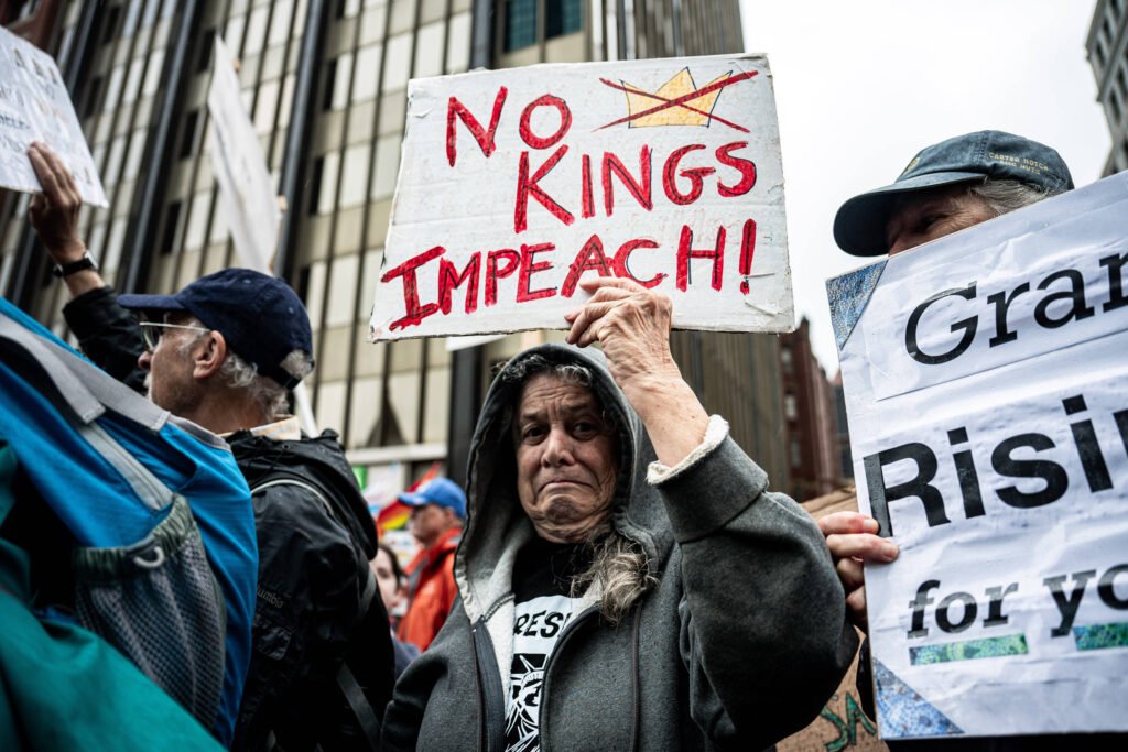 Manifestante morre baleado em manifestação contra Trump A imagem mostra um grupo de manifestantes em um protesto. Em primeiro plano, uma mulher com cabelo grisalho segura um cartaz que diz