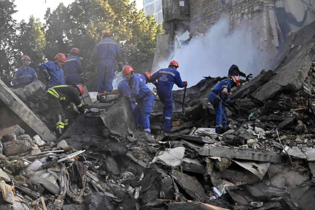 Um grupo de pessoas, incluindo bombeiros e socorristas, está trabalhando em meio aos escombros de um edifício desabado. Eles usam capacetes e roupas de proteção, e alguns estão utilizando ferramentas para remover os destroços. Há fumaça visível no fundo, indicando uma possível situação de emergência. A cena é de intensa atividade de resgate.