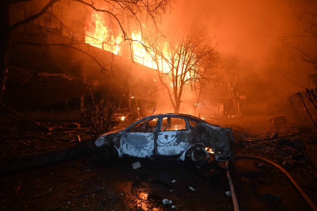 A imagem mostra um carro danificado em primeiro plano, com a carroceria queimada e em estado de abandono. Ao fundo, há um grande incêndio, com chamas intensas e fumaça, iluminando a cena com uma luz laranja. Árvores estão visíveis, algumas delas também afetadas pelo fogo. O ambiente parece caótico e devastado, sugerindo um incêndio em uma área urbana.