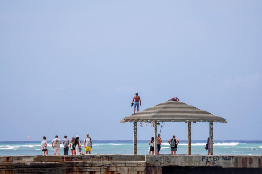A imagem mostra um grupo de pessoas em um píer, algumas delas em pé e outras sentadas. No fundo, há um homem em cima de uma estrutura coberta, observando o mar. O céu está claro e há ondas visíveis no oceano. O píer parece ser de madeira e há grafites visíveis na estrutura.