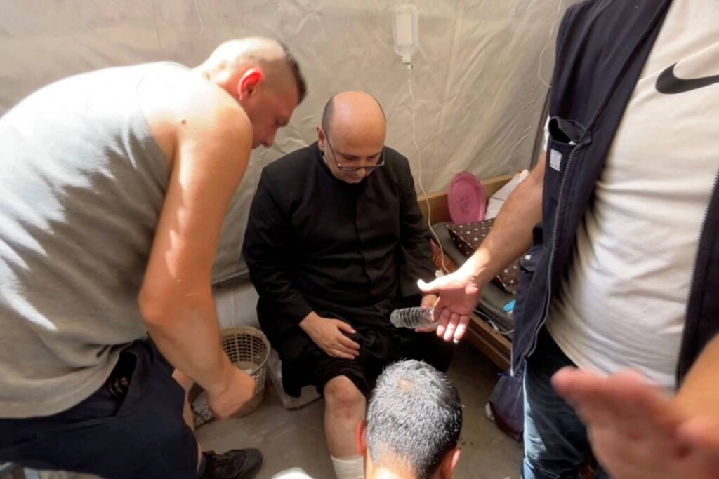 A imagem mostra um grupo de homens em um ambiente interno. Um homem, vestido com uma roupa escura, está sentado e parece estar recebendo cuidados ou assistência de outros homens ao seu redor. Um deles está agachado perto de seus pés, enquanto os outros estão em pé, observando ou ajudando. O ambiente parece ser simples, com paredes claras e alguns objetos ao fundo.