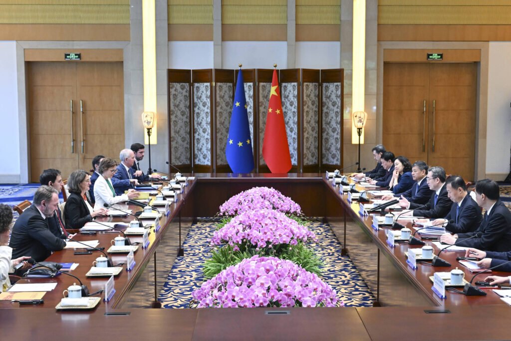A imagem mostra uma sala de conferências onde ocorre uma reunião entre representantes da União Europeia e da China. No centro da mesa, há um arranjo de flores rosas. Ao fundo, estão as bandeiras da União Europeia e da China. Os participantes estão sentados em ambos os lados da mesa, com documentos e copos à frente.