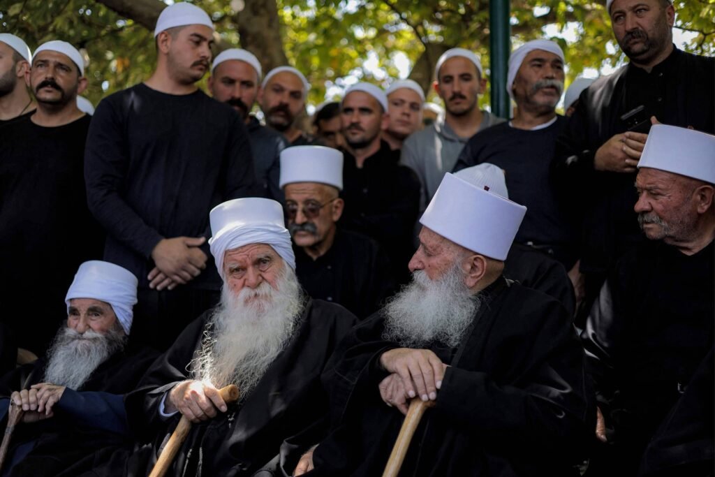 A imagem mostra um grupo de homens, muitos deles com turbantes brancos, sentados e em pé. Os homens mais velhos estão usando roupas escuras e têm longas barbas brancas. Eles parecem estar em um ambiente ao ar livre, com árvores ao fundo. O grupo inclui tanto homens mais velhos quanto mais jovens, todos com expressões sérias.