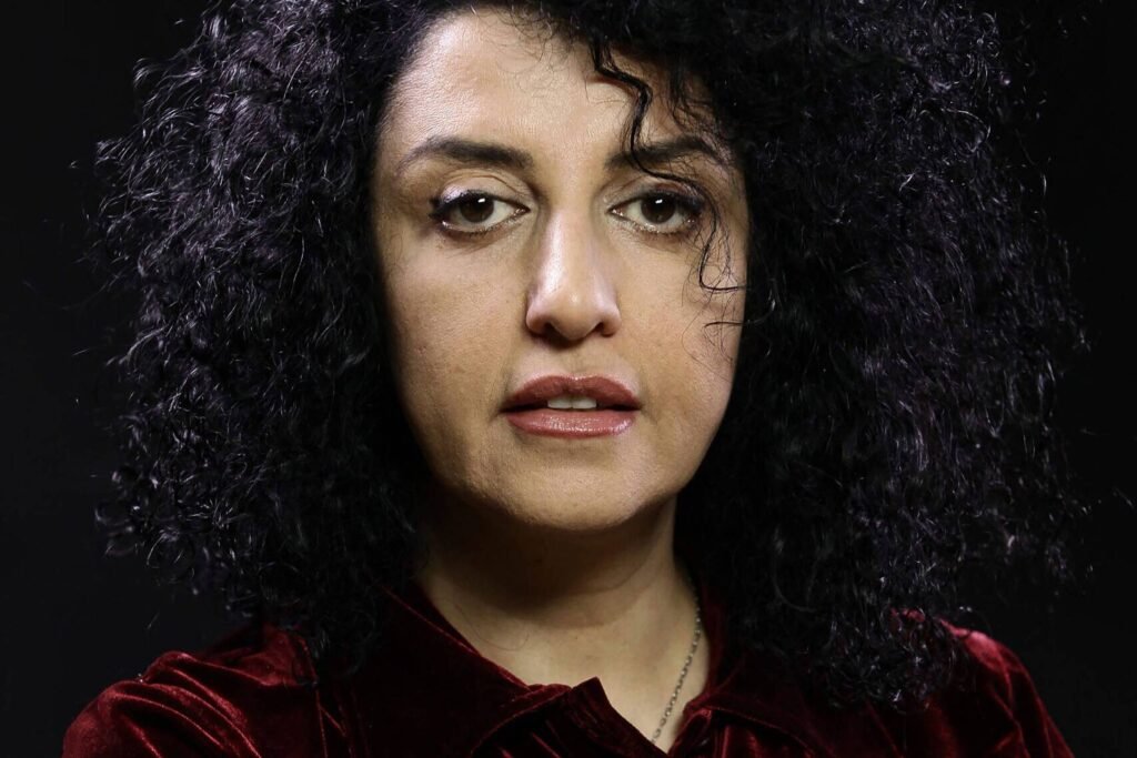A imagem mostra uma mulher com cabelo cacheado e volumoso, usando uma blusa de veludo vermelho. Ela está olhando diretamente para a câmera com uma expressão séria. O fundo é escuro, destacando seu rosto e a textura do cabelo.