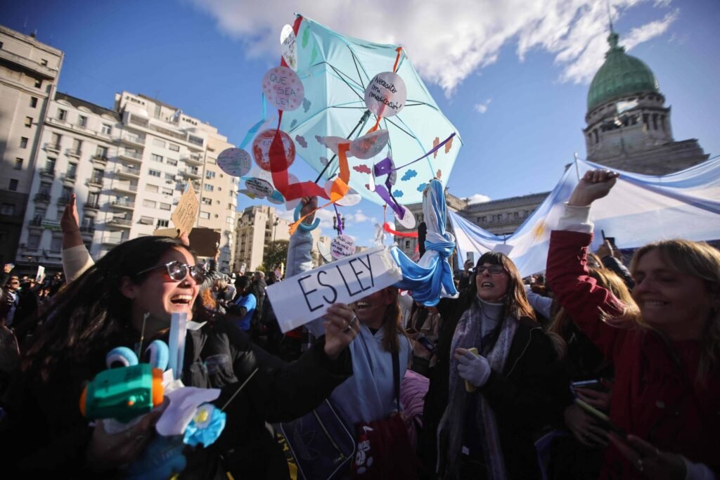 A imagem mostra uma manifestação em um espaço público, onde várias pessoas estão reunidas. Algumas delas seguram um guarda-chuva colorido decorado com objetos e desenhos. Uma mulher à frente segura um cartaz com o nome