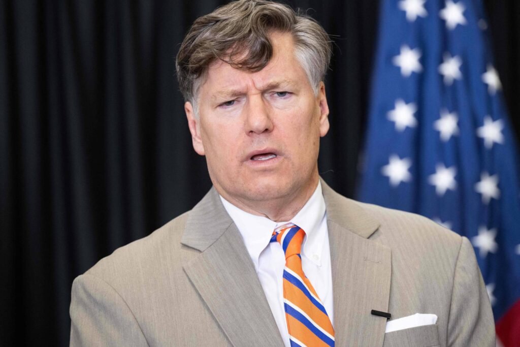 Um homem com cabelo castanho e um terno claro, usando uma gravata listrada em azul e laranja. Ele parece estar em uma conferência ou evento, com uma expressão facial séria. Ao fundo, há uma bandeira com estrelas e listras, sugerindo um ambiente formal ou governamental.