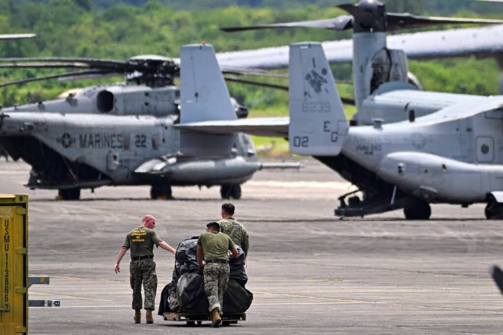 A imagem mostra um cenário militar com dois helicópteros de transporte, identificados como pertencentes aos fuzileiros navais dos EUA, estacionados em uma pista. Três militares estão empurrando um carrinho com um objeto coberto por uma lona, enquanto um dos helicópteros está visível ao fundo. O ambiente é ao ar livre, com vegetação ao fundo e um céu claro.